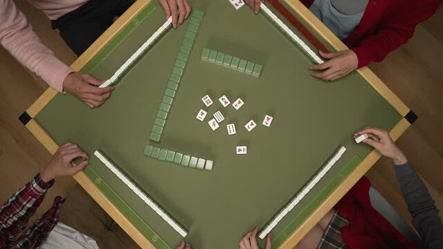view from above of people's hands picking and laying down green mahjong pieces on the table while playing tile game together at home during chinese new year holiday