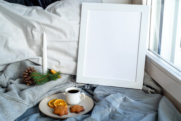 Cozy Christmas breakfast still life scene near the window. Winter interior.Blank white picture frame mockup.Cup of coffee, wholemeal cookies, pillow, wool sweater, candle. Hotel breakfast menu templat