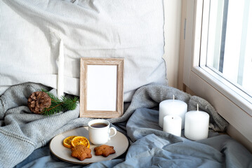 Cozy Christmas breakfast still life scene near the window. Winter interior.Blank wooden picture frame mockup.Cup of coffee, wholemeal cookies, pillow, wool sweater,candle.Hotel breakfast menu template