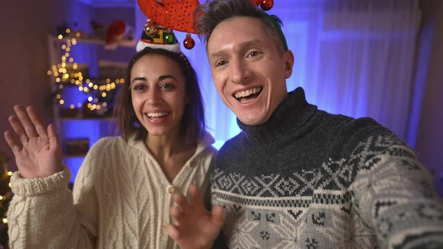 POV first person view happy cheerful excited couple, woman in festive Elf Cap and man in red reindeer antlers headband taking selfie on background of decorated house with christmas tree