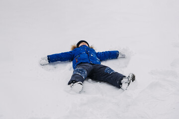 Fototapeta na wymiar Best Outdoor Winter Activities for kids. Little kid boy in blue winter jacket making snow angel, laying down on snow in winter backyard. Active outdoors leisure with children in winter.