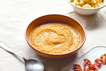 Pumpkin cream soup with roasted bacon and croutons in clay plate on the table with linen tablecloth. Autumn vegetable creamy soup puree. Healthy seasonal eating.