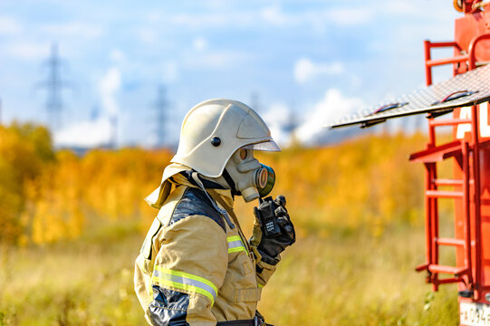 A firefighter rescuer in a protective suit, gas mask and white helmet extinguishes a fire at a metallurgical plant, keeps in touch by talking on a walkie-talkie. Fire extinguishing operation. .