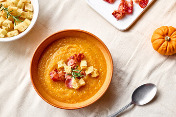 Pumpkin cream soup with roasted bacon and croutons in clay plate on the table with linen tablecloth. Autumn vegetable creamy soup puree. Healthy seasonal eating.