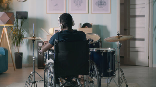 Back view of anonymous black man with disability sitting on wheelchair and playing drums during rehearsal in light home studio in daytime