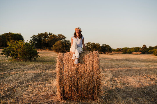 Woman wearing hat sitting on straw bale during sunset