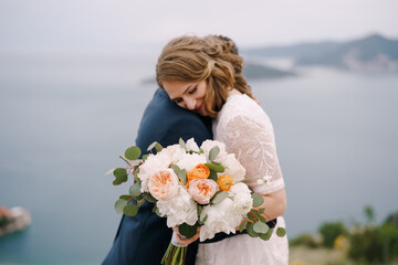 Groom hugs bride with a bouquet of flowers while standing on the mountain above the sea. Portrait