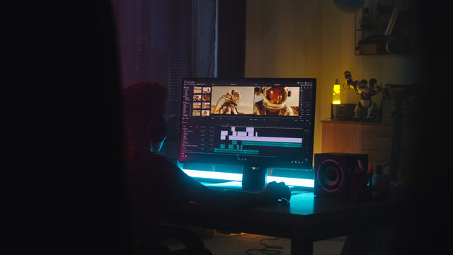 Teenager using computer to edit footage while sitting at desk in obscure room at night at home
