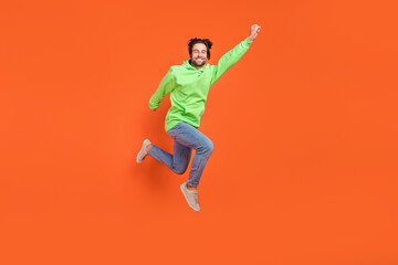 Fototapeta na wymiar Full size photo of brunet young guy jump save world wear sweater jeans sneakers isolated on orange background