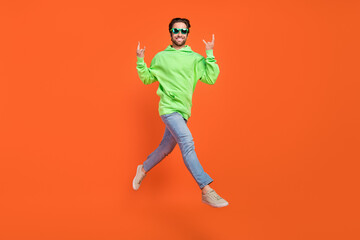 Fototapeta na wymiar Full size photo of brunet young guy run show rock sign wear hoodie jeans sneakers eyewear isolated on orange background