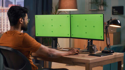 Bearded Indian guy in glasses sitting at table and browsing data on computer with chromakey...