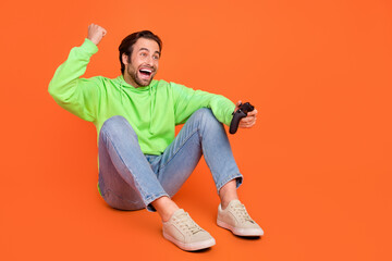 Full length photo of brunet young guy play station sit look promo wear green sport cloth jeans footwear isolated on orange color background