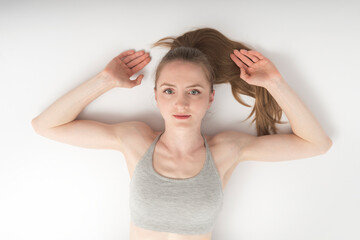 Naklejka premium Beautiful girl lies on white floor and raised hands up. Portrait of young woman in sports top on white background. Top view