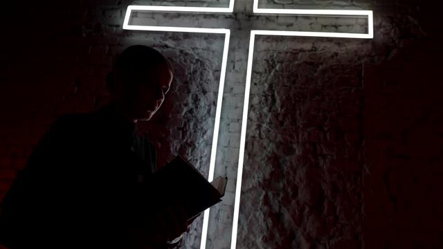 a woman in black clothes stands under a glowing cross in the dark and reads a text from a book