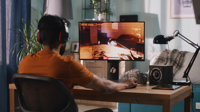 Side view of bearded Indian man in casual clothes and wireless headphones sitting at table and playing shooter on computer in weekend at home