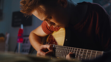 Low angle of concentrated teenager with pick practicing to play electric guitar while sitting on...