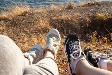 Two pairs of female legs in sports sneakers on the seashore. Female feet seen from above. Girls sitting on a hill overlooking the ocean - Powered by Adobe