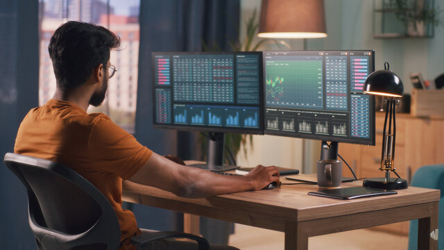 Smart bearded Indian broker sitting at table and using computer to trade on stock market during work in evening at home