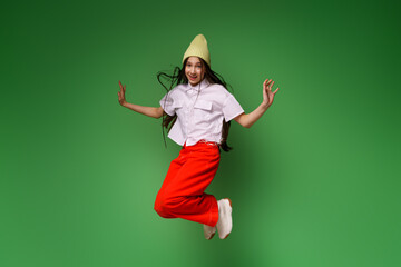 Full length photo of positive cheerful teenage girl jumping up in white shirt and cap, red pants and white sneakers, isolated on green colored background