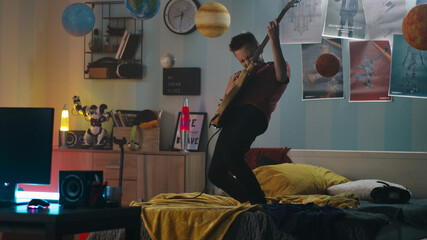 Zoom in view of teen musician playing energetic song on electric guitar while standing on bed near...