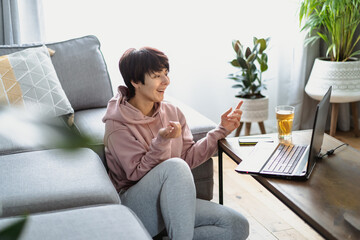 Emotional Woman in casual home clothes having video call sitting near sofa using a laptop. Cozy home workspace in modern interior with home plants. Remote work from home. Selective focus, copy space.
