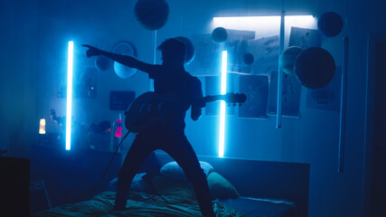 Pan around view of male teenager in glowing sunglasses playing loud music on electric guitar while standing on bed against blue neon lamps and he imagines that he is a rockstar on stage