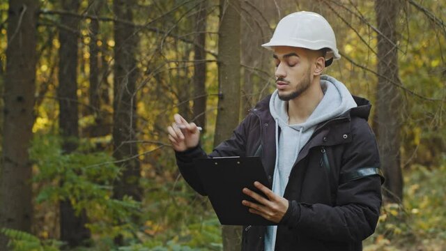 Forest evaluation and management. Millennial Indian Forestry engineer in hardhat with tablet in woods. Forester foreman working and supervising in park, watching of wildlife sanctuary checking trees