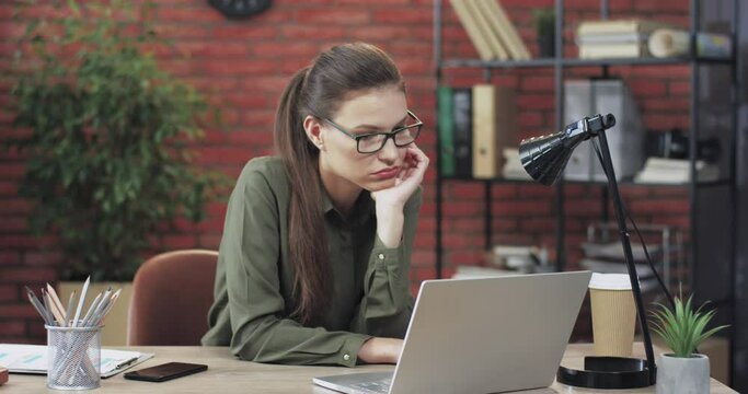 Young beautiful caucasian woman with glasses sitting in modern office looking upset at laptop, tired of unresolved task.
