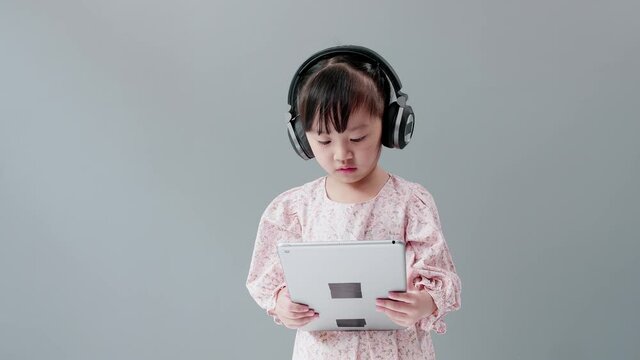 ittle Asian girl with headphone enjoy watching video online on tablet and dancing happily.