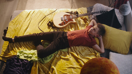From above teenager with hands behind head lying on bed near electric guitar and resting under planets models during rehearsal in bedroom at home