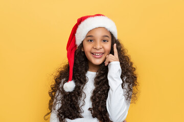 Studio shot of dark skinned young girl with long curly hair in a Santa Claus hat on a yellow background