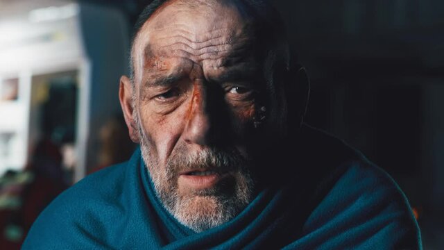 Unhappy aged man with dirty face wrapping in blanket and looking at camera after accident at dark knight