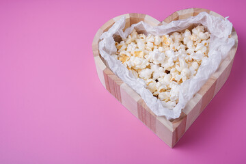 Wooden heart shaped box with popcorn on pink background, top view, copy space. Close up shot