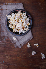 Heap of popcorn in black bowl on wooden rustic background. Some popcorns on fabric and table. Vertical shot, top view