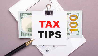 On a gray background, a white calculator, a pen, banknotes and a sheet of paper under a black paper clip with the text TAX TIPS. Business concept