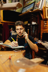 Young man in his craft workshop talking on the phone, picking up orders and writing down a notebook.