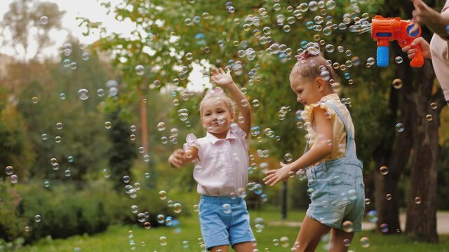 Happy mother blowing soap bubbles with toy gun, little daughters trying to catch bubbles flying in air. Family playing together, have fun in park. Mother's Day Joy, Mom and Baby
