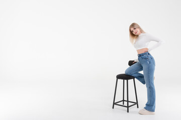 Happy and beautiful young woman in a white sweater and jeans with a black bar stool on a white background. People lifestyle concept.