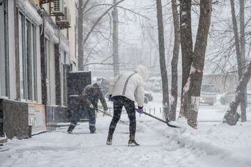 A girl dressed in light winter clothes is shoveling snow off the walkway. Behind her is a man with a shovel.
