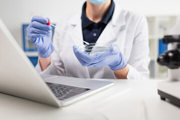 Cropped view of scientist holding petri dish and pcr test near laptop in lab.