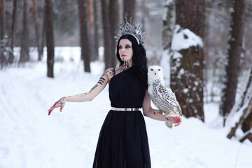 Beautiful brunette woman with body art in crown and black long dress with polar owl on hand standing in winter forest © Альбина Хусаинова