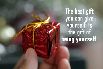 Inspirational quote - The best gift you can give yourself, is the gift of being yourself. Person holding red Christmas box in hand on soft colorful bur bokeh background. Self love and care concept.
