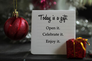 Life inspirational quote on a notepaper - Today is a gift. Open it. Celebrate it. Enjoy it. With red Christmas ball ornament  and red gift box decoration on gray silver color background.