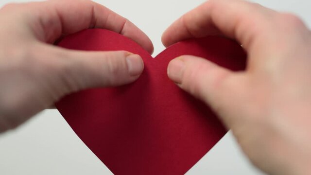 hands tearing big red paper heart on two half halves, throwing on white table surface. close up view. broken love, abusive relationship, end of marriage concept. pain from breaking up divorce problem