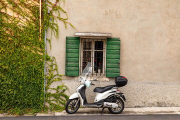Typical Italian street scene with a white scooter by a window with green shutters on a tarmac...