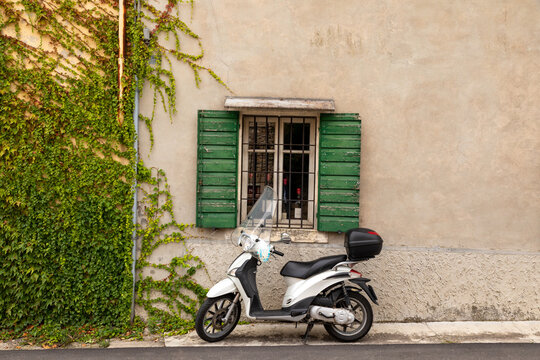 Typical Italian street scene with a white scooter by a window with green shutters on a tarmac street in Colognola ai Colli, Verona, Italy.