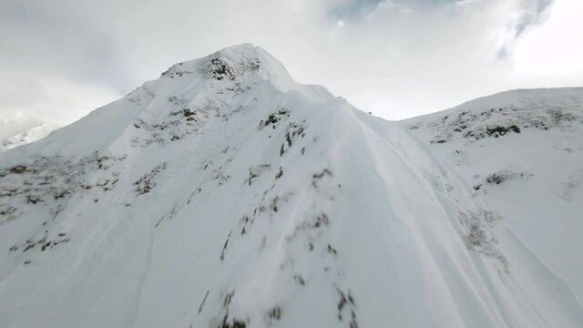 Aerial view sports FPV drone flying up mountain ridge with people riding from top on snowboard enjoying extreme activity hobby. Amazing wild nature landscape winter panorama with athletic tourists