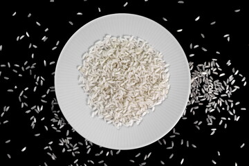 Rice in a white plate and grains spilled on a black background.