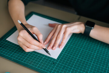 Close-up top view of unrecognizable female designer drawing making mark with pencil on white blank paper lying on green rubber cutting mat at desk, selective focus. Concept of creative work and hobby