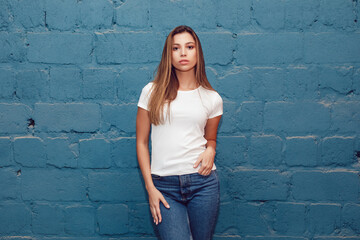 Fototapeta na wymiar Beautiful young woman in white blank t-shirt stands against a blue brick wall background.
